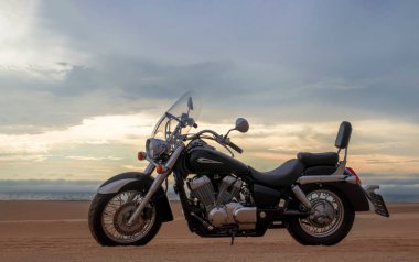 Namib Çölü 'nün kumlu yüzeyinde dramatik bulutlu bir gökyüzüne karşı tek başına duran bir motosiklet. Günbatımı ışığı, doğayla makine arasındaki zıtlığı vurgular..