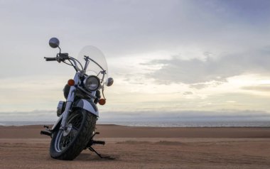Namib Çölü 'nün kumlu yüzeyinde dramatik bulutlu bir gökyüzüne karşı tek başına duran bir motosiklet. Günbatımı ışığı, doğayla makine arasındaki zıtlığı vurgular..