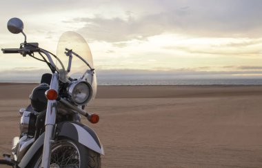 Namib Çölü 'nün kumlu yüzeyinde dramatik bulutlu bir gökyüzüne karşı tek başına duran bir motosiklet. Günbatımı ışığı, doğayla makine arasındaki zıtlığı vurgular..