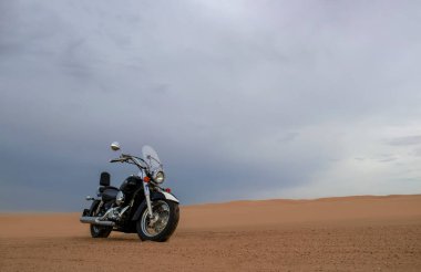 Namib Çölü 'nün kumlu yüzeyinde dramatik bulutlu bir gökyüzüne karşı tek başına duran bir motosiklet. Günbatımı ışığı, doğayla makine arasındaki zıtlığı vurgular..