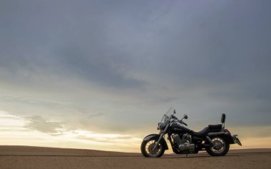 Namib Çölü 'nün kumlu yüzeyinde dramatik bulutlu bir gökyüzüne karşı tek başına duran bir motosiklet. Günbatımı ışığı, doğayla makine arasındaki zıtlığı vurgular..