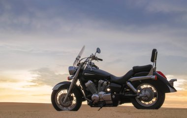 Namib Çölü 'nün kumlu yüzeyinde dramatik bulutlu bir gökyüzüne karşı tek başına duran bir motosiklet. Günbatımı ışığı, doğayla makine arasındaki zıtlığı vurgular..