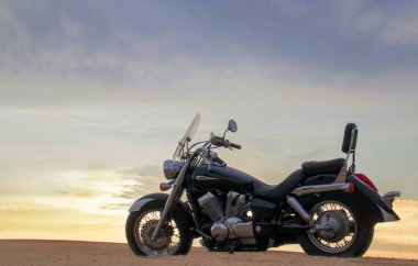 Namib Çölü 'nün kumlu yüzeyinde dramatik bulutlu bir gökyüzüne karşı tek başına duran bir motosiklet. Günbatımı ışığı, doğayla makine arasındaki zıtlığı vurgular..