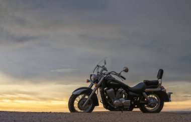 Bir motosiklet, gün batımının altında hareketsiz durur. Özgürlüğü, macerayı ve önümüzde yeni yollar keşfetmenin heyecanını sembolize eder..