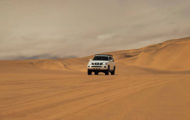 Beyaz 4x4 SUV Namib Çölü 'nde altın kum tepelerini aşıyor. Hareketli bir off-road aracı, dramatik bir çöl zeminine doğru parlak güneş ışığında yakalandı.