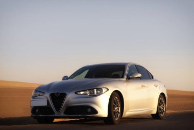 Lüks Alfa Romeo Giulia Namibya çölünde dinleniyor, kum tepeleri ve kuru arazi ile çevrili. Editör araba fotoğrafçılığı tasarım ve doğa arasındaki zıtlığı yakalıyor. 