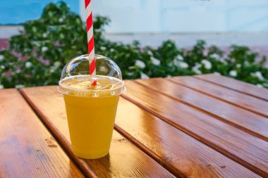 Plastik bardak taze sıkılmış organik portakal suyu. Dışarıdaki tahta masada kapağı ve samanı var. Lezzetli ve sağlıklı bir içecek. Antioksidan diyet içeceği.