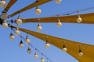 Tente desenli üçgen kumaş esneme çatısı. Mavi gökyüzü arka planında parti gecesi ışıklandırması için sıra sıra asılı ampullerle yelken açıyor. Açık hava lamba telleri ve naylon güneş gölgeleri