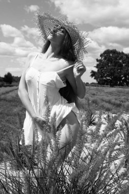 Hasır şapkalı seksi bir kadın bahçede gökyüzünün arka planında yürüyor. Siyah beyaz fotoğraf..