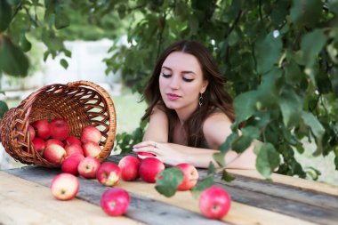 Genç ve güzel bir kadın elma bahçesinde bir sepet ve dağınık meyveyle oturuyor..