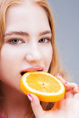 Gri arka planda izole edilmiş olgun, sulu bir portakal yiyen güzel bir kadının portresi. Sağlıklı beslenme konsepti. Diyet.