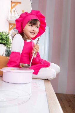 Aşçı kostümü giymiş küçük güzel kız mutfakta fırının yanında otururken yemek hazırlıyor..