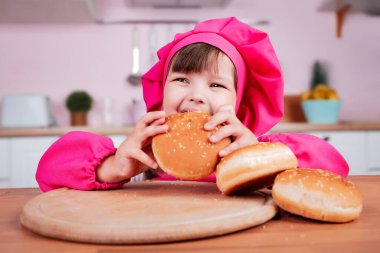 Mutfak masasında otururken aşçı kostümü giymiş küçük mutlu kız çörekleri yiyor. Sağlıklı beslenme.