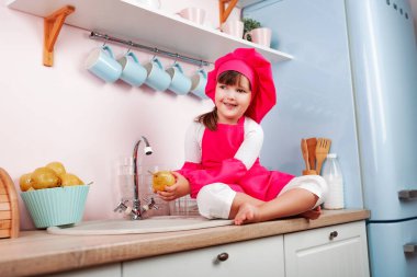 Aşçı kostümü giymiş mutlu küçük güzel kız mutfaktaki fırının yanında otururken musluğun altında armut yıkıyor..