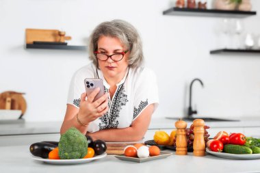 Mutfakta yemek hazırlarken akıllı telefona bakan güzel bir kadın. Sağlıklı beslenme. Kilo verme konsepti. Vitamin gıdaları sağlığınız için iyidir. İnternetteki tarifler. Blogcu.