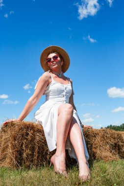 Hasır şapkalı ve güneş gözlüklü genç ve güzel bir kadın güneşli bir günde gökyüzünde samanlıkta oturuyor. Dinlen. Tatilde. Turizm.
