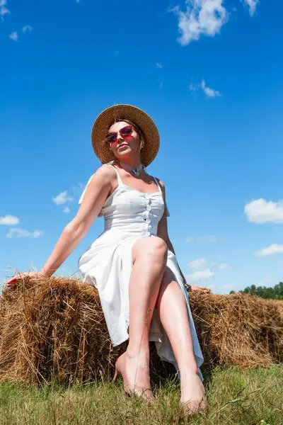 Hasır şapkalı ve güneş gözlüklü genç ve güzel bir kadın güneşli bir günde gökyüzünde samanlıkta oturuyor. Dinlen. Tatilde. Turizm.