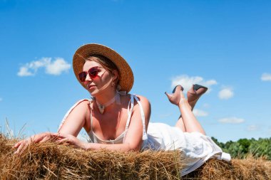 Hasır şapkalı ve güneş gözlüklü genç ve güzel bir kadın güneşli bir günde samanlıkta gökyüzüne karşı yatıyor. Dinlen. Tatilde. Turizm. Romantizm. Kırsal.