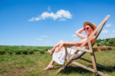 Hasır şapkalı ve güneş gözlüklü güzel bir kadın güneşli bir günde güneşli bir çayırda güneşli bir yerde oturuyor. Dinlen. Tatilde. Turizm.