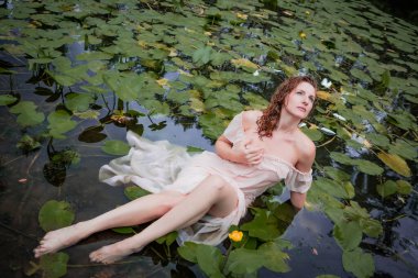 Gölde çiçekli nilüferler içinde elbisesiyle yatan seksi, ıslak, genç bir kadın. Romantizm, rahatla, turizm. Üst görünüm.
