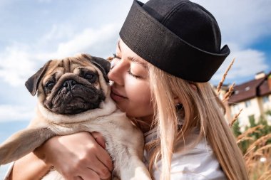 Genç ve mutlu bir kadın şehir parkında koşum takımı ve siyah tasmayla yürürken sevgili köpeğini tutuyor, öpüyor ve sarılıyor. Sokak fotoğrafı..
