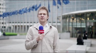 Bir TV haber yayınının son dakika gelişmesiyle birlikte arka planda mavi bayraklar sallanan modern bir kurumsal binanın dışından bir gazeteci canlı yayın yapıyor.