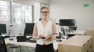Çağdaş bir ofiste, kendine güvenen genç profesyonel bir kadın elinde bilgisayarlı belgeler tutarken görülüyor, yenilik ve takım çalışması ile dolu bir şirket ortamını yansıtıyor.