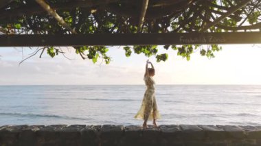 Zarif çiçekli bir elbise giymiş şık bir bayan, yapraklı bir tepe örtüsünün altında huzurlu bir gezinti yapıyor. Huzurlu bir okyanus ve ılık bir günbatımı manzaralı bir zemin oluşturuyor.