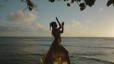 Gün batımında okyanus kenarında geleneksel hareketler yapan bir dansçının güzel görüntüsünün tadını çıkarın. Arka planda sakin dalgalar ve çarpıcı bir siluet yaratan güzel bir gökyüzü var.