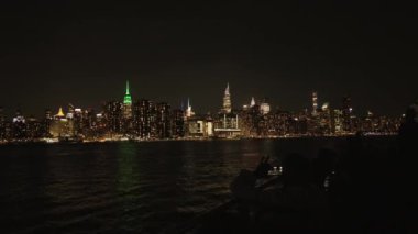 New York City 'nin ikonik gökdeleninin nefes kesici akşam manzarası. Empire State Binası gibi parlayan gökdelenler aşağıdaki nehri yansıtıyor. Şehirlerin mimari güzelliğini gözler önüne seriyor.