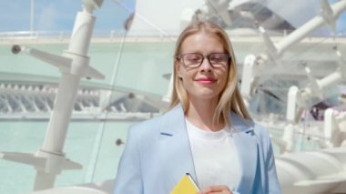 Açık mavi ceketli kendine güvenen bir iş kadını elinde bir defter tutuyor, modern şehir mimarisinin önünde duruyor ve arkasında parlak bir gökyüzü var. Profesyonellik ve liderlik yayıyor.