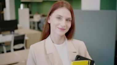 Profesyonel kıyafetli, kendine güveni olan genç bir işçi modern bir ofiste defterlerle duruyor. Çeşitli meslektaşlar, takım çalışmasını ve başarıyı sembolize ederek arka planda işbirliği yaparlar.