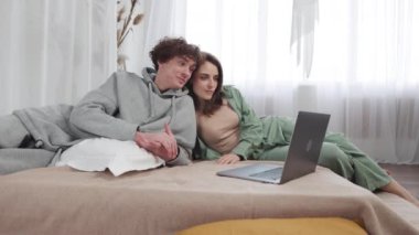 Bir çift dizüstü bilgisayarda bir video izliyor, yatakta gülümsüyor ve rahatlıyor, pozitif ve sevgi dolu bir bağ gösteriyor.