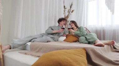Rahat giysiler içinde genç bir çift yatak odalarında şarabın tadını çıkarıyorlar, evlerinde rahat ve sevgi dolu bir atmosferde, kişisel dokunuşlar ve yumuşak ışıklandırmayla çevrili özel anları paylaşıyorlar.