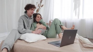 Genç bir çift, rahat giysiler içinde yataklarında rahat bir şekilde rahat bir şekilde dizüstü bilgisayardan parlak ve sıcak bir ev ortamında bir şeyler izliyor. Sahne samimi ve samimi.