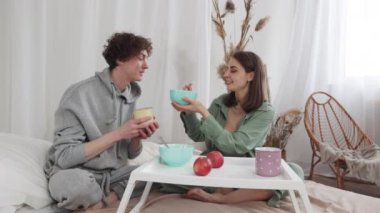 Neşeli bir çift yatakta kahvaltı yapar, güler ve birbirlerini beslerler. Sevgi dolu ve samimi bir sabah anı.