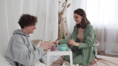 Genç bir çift yatakta rahat bir anı paylaşıyor, kahve içiyor ve rahat bir yatak odası ortamında konuşuyor..