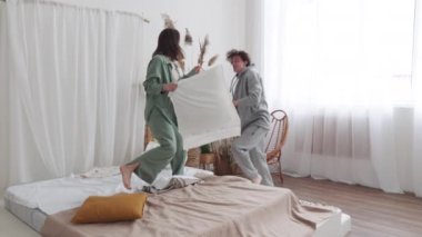 Genç çift, rahat, modern bir yatak odasında beyaz perdeleri ve doğal dekorları olan bir yastık kavgasının keyfini çıkarıyor..