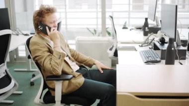 Modern bir ofiste genç bir profesyonel, bir telefon görüşmesi yapıyor, kurumsal çevrenin dinamiklerini sergiliyor. Anahtar kelimeler iş, iletişim, etkileşim, takım çalışması, teknoloji