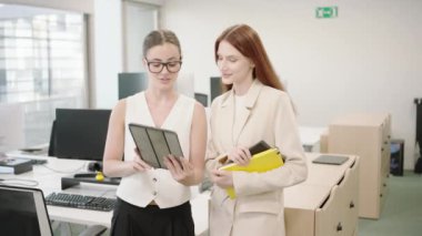 Modern bir ofiste yapılan bir iş toplantısında, iki profesyonel kadın işbirliği yapmak ve belgeleri gözden geçirmek için bir tablet kullanıyor. Arka planda çeşitli bilgisayar ve ofis ekipmanları görünür