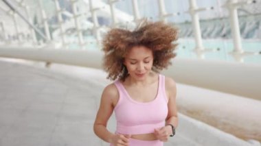 Pembe spor giyimli dinamik genç bir kadın canlı bir şehir ortamında arka planda modern mimari ile koşuyor, enerjik yaşam tarzını ve fitness 'e bağlılığını gösteriyor.