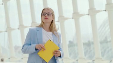 Ağır çekimde, bu sahnede zarif bir iş kadını mavi ceketli, zarif bir şekilde sarı bir klasörü tutarken sofistike bir marina ortamında yürüyor. Yavaş Hareket 