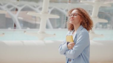 Kıvırcık saçlı ve gözlüklü genç ve güzel bir kadın, açık ve modern bir ortamda sarı bir defter tutarken gülümsüyor. Mutluluk ve memnuniyet saçar, kendinden emin, profesyonel görünür.,