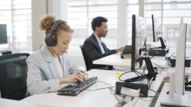 Modern bir ofiste özenle çalışan başarılı genç profesyoneller. Kulaklık takan bir kadın, arka planda çalışan bir erkek. Sahne takım çalışmasını ve üretkenliği tasvir ediyor.