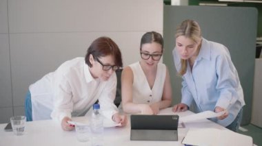 Çağdaş bir ofis ortamında, üç genç iş kadını bir tablet üzerindeki verileri inceliyor, bir proje üzerinde işbirliği yapıyor ve belgeleri inceliyor, takım çalışması ve adanmışlığı gösteriyor
