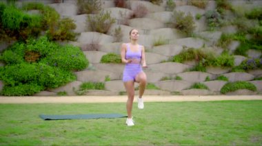 Güzel bir kadın zarif bir şekilde yoga minderinin üzerinde açık hava ortamında çeşitli spor egzersizleri yapıyor. Koşarak spor ve sağlığa olan bağlılığını etkili bir şekilde gösteriyor.
