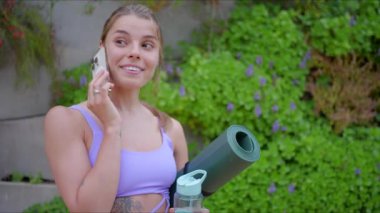 Genç, zinde bir kadın coşkuyla bir yoga minderi ve su şişesi ile enerji dolu bir egzersiz seansına hazırlanıyor. Atletizmini ve sağlıklı yaşam tarzını mükemmel bir şekilde gösteriyor.