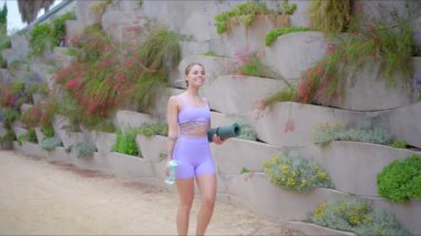 Havalı atletik giyinen güçlü bir kadın, canlı bir açık hava ortamında koşar, yoga minderi ve su şişesi taşır, spor ve sağlığa bağlılığını gösterir.