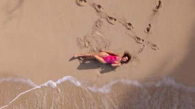 Gündoğumunda Oahu 'da sakin bir plajın huzurlu güzelliğindeki kadınları çeken bir hava çekimi, canlı pembe mayo giymiş bir figürü hafif dalgalara ve yumuşak, davetkar kuma karşı çeker. Hawaii