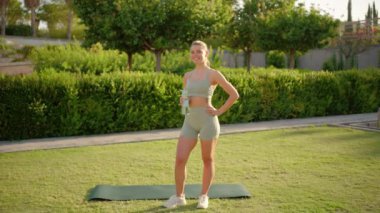 Neşeli ve hayat dolu genç bir kadın, doğa tarafından çevrelenmiş, güzel ve bereketli bir açık hava ortamında yoga minderinde pozlar vererek çeşitli fitness aktivitelerine katılıyor.